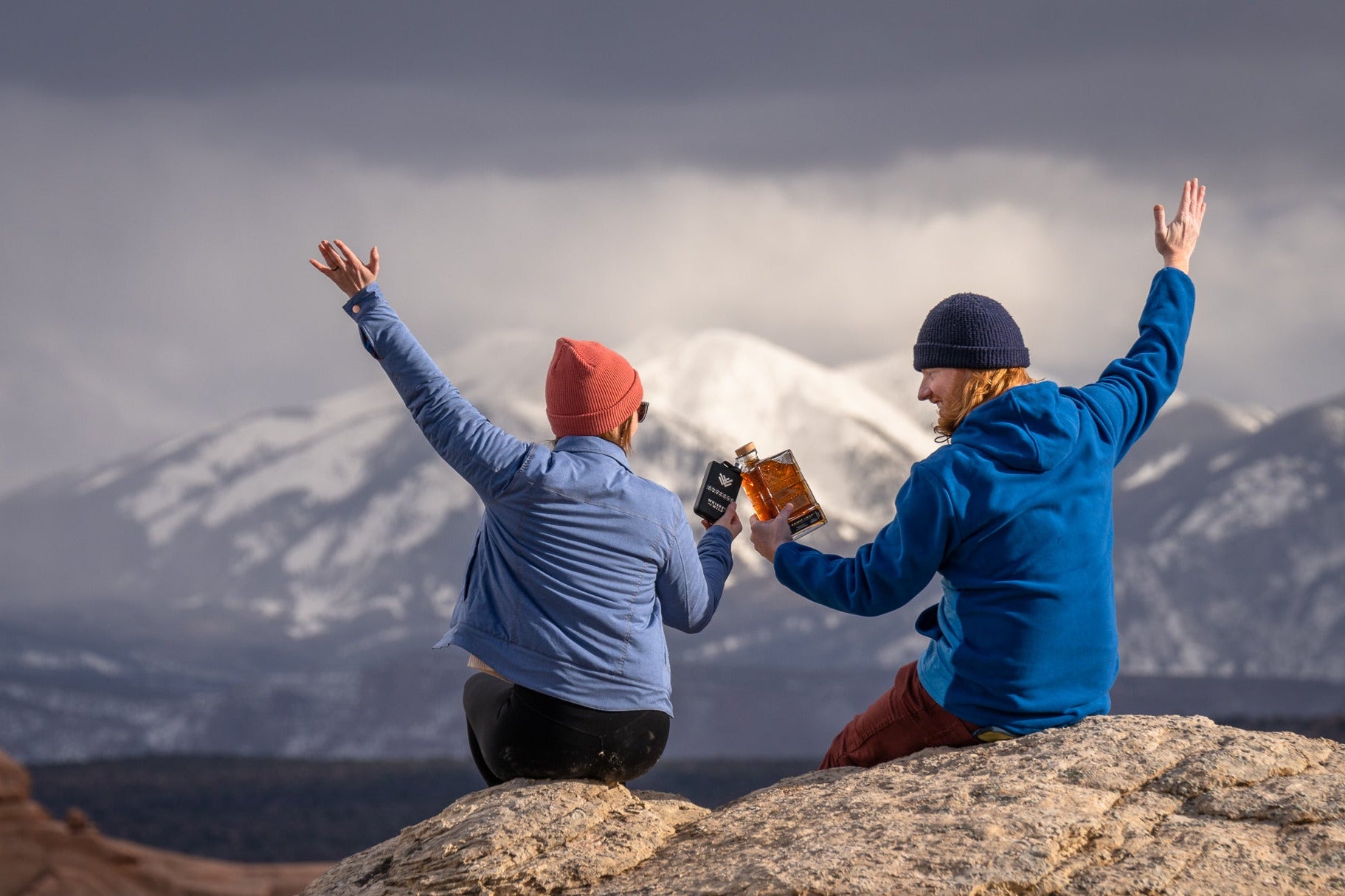 Two people sitting on a rocky outcrop with arms raised, enjoying a scenic view of mountains while holding a bottle and flask from Whiskey in the Wild.